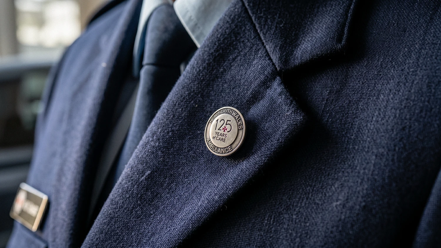A silver 125 Years of Care commemorative pin on a navy ambulance uniform lapel
