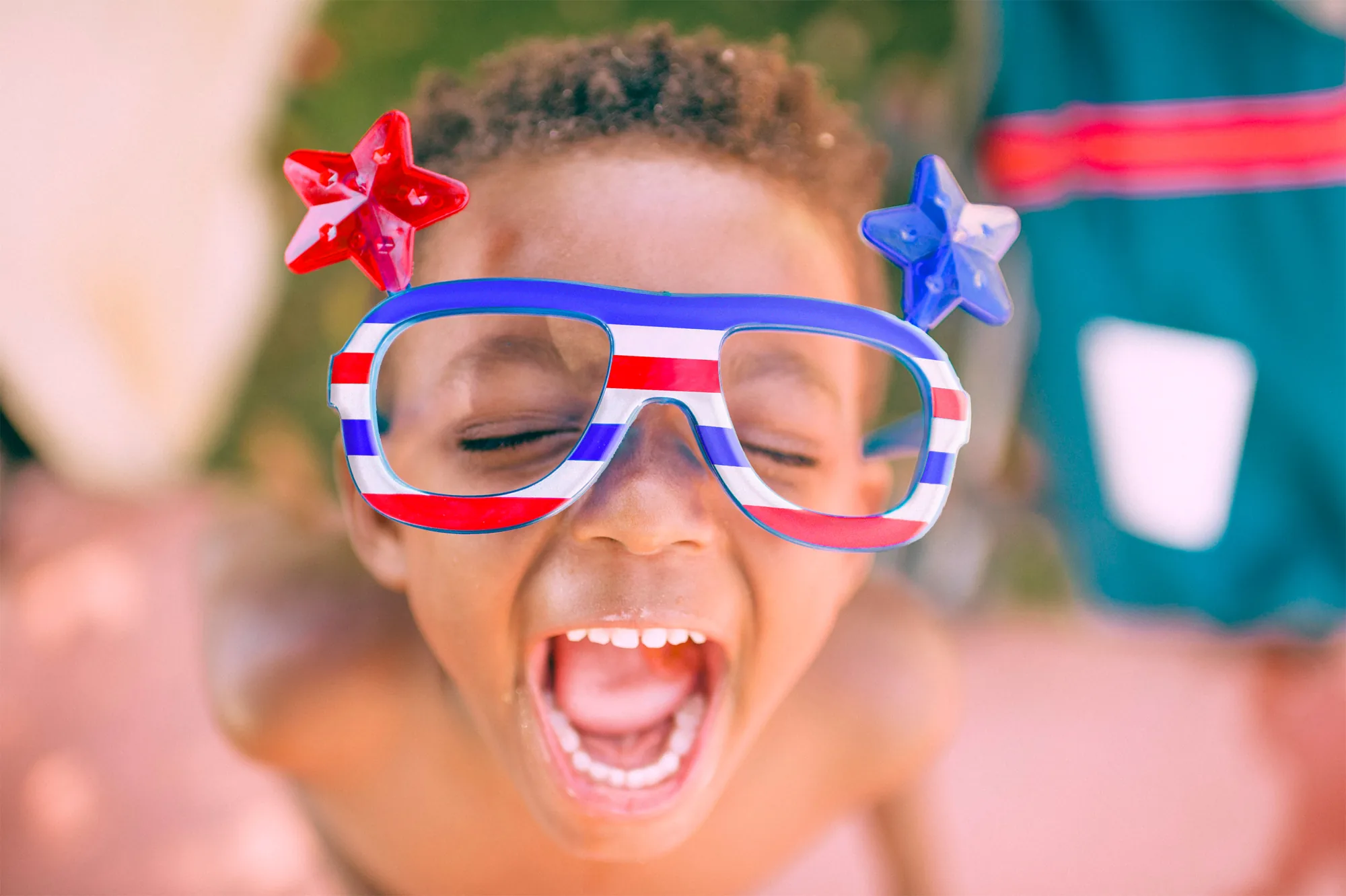 A laughing child outdoors wearing star-topped red, white and blue striped novelty glasses.