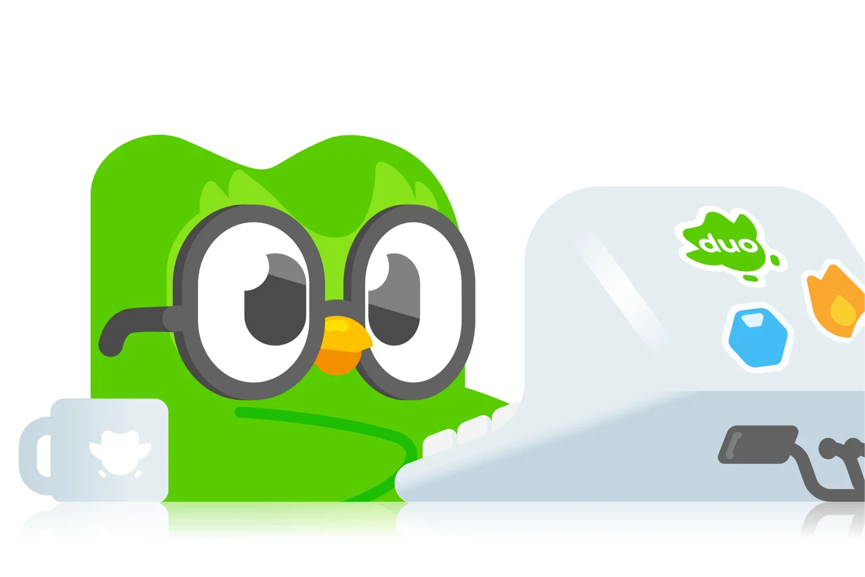 Duolingo's owl mascot Duo peering over a laptop covered in brand stickers